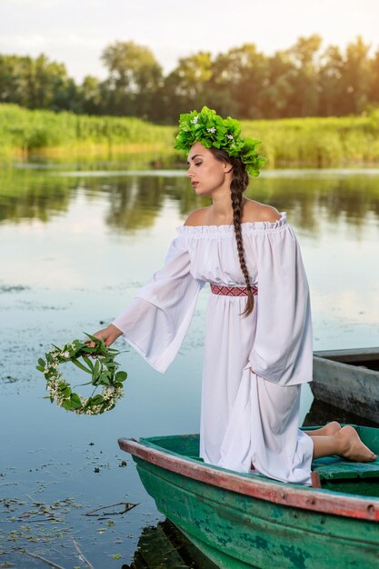 Junge sexy Frau auf Boot bei Sonnenuntergang. Das Mädchen hat einen Blumenkranz auf dem Kopf, entspannt sich und segelt auf dem Fluss. Schöner Körper und Gesicht. Fantasy-Kunstfotografie. Konzept der weiblichen Schönheit, Ruhe im Bösen