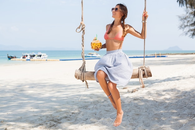 Junge sexy Frau sitzt auf Schaukel am tropischen Strand, Sommerferien, Modestil, Rock, Bikinioberteil, trinkenden Kokosnusscocktail, lächelnd, entspannend