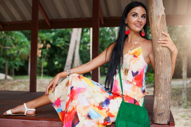 Junge sexy schöne Frau im bunten Kleid, Sommer Hippie-Stil, tropischen Urlaub, gebräunte Beine, Sandalen, grüne Handtasche mit Fransen, Accessoires, lächelnd, glücklich