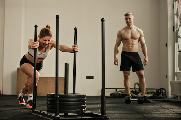 Junge Sportlerin macht Schlittenschubübungen und schreit während des Sporttrainings in einem Fitnessstudio, während ihre Freundin sie beobachtet