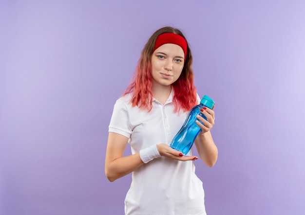 Junge sportliche Frau, die Flasche Wasser mit sicherem Lächeln hält, das über lila Wand steht