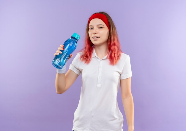 Kostenloses Foto junge sportliche frau, die flasche wasser mit sicherem lächeln hält, das über lila wand steht