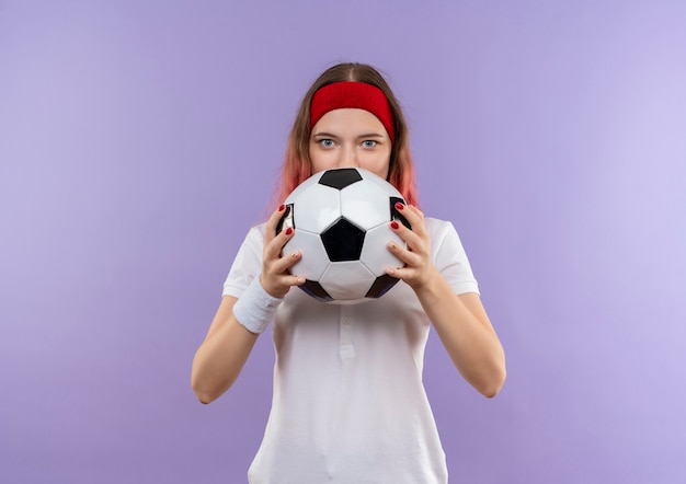 Junge sportliche Frau, die Fußball hält, der ihr Gesicht hinter Ball hält, der über lila Wand steht