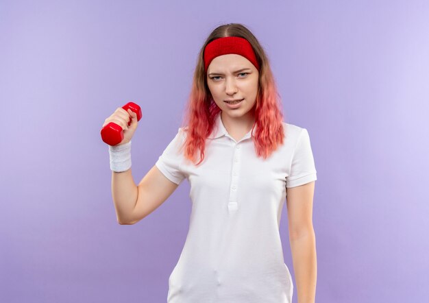 Junge sportliche Frau, die Hantel in der Hand mit ernstem Gesicht hält, das über lila Wand steht