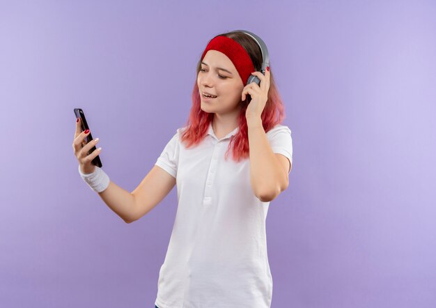 Junge sportliche Frau, die Musik mit ihren Kopfhörern betrachtet, die Bildschirm ihres Smartphones lächelnd stehen über lila Wand betrachten