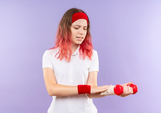 Junge sportliche Frau, die zwei Hanteln hält, die Übungen betrachten, die ihr verbundenes Handgelenk betrachten, das Schmerz empfindet, der über lila Wand steht