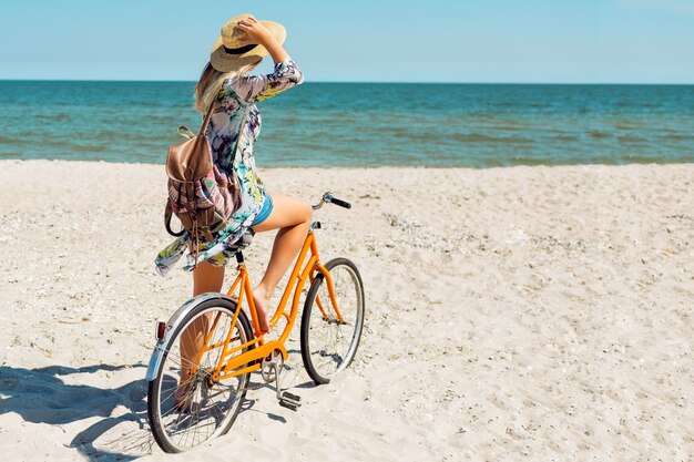 Junge sportliche Frau im stilvollen weißen Erntedach und in den Jeansshorts, die am Strand mit orange Fahrrad stehen