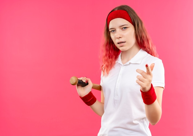 Junge sportliche Frau im Stirnband, die Baseballschläger hält, der zuversichtlich zeigt, mit dem Finger zur Kamera über rosa Wand stehend
