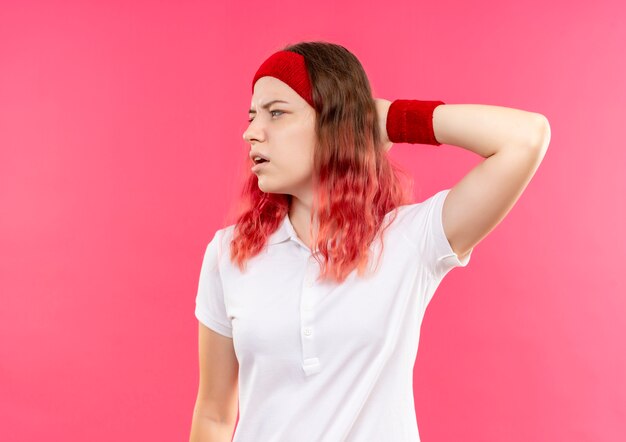 Junge sportliche Frau im Stirnband, die beiseite mit nachdenklichem Ausdruck steht, der über rosa Wand steht