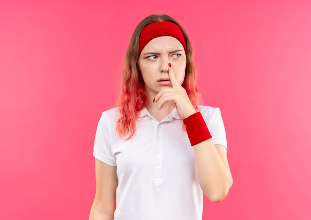 Junge sportliche Frau im Stirnband, die beiseite schaut, berührt ihre Nase mit nachdenklichem Ausdruck auf Gesicht, das über rosa Wand steht
