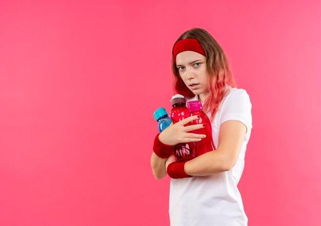 Junge sportliche Frau im Stirnband, die Flaschen des Wassers mit dem Angstausdruck über rosa Wand umarmt