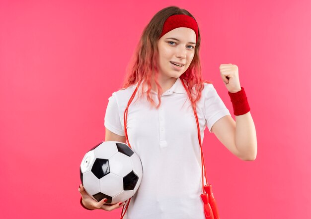 Junge sportliche Frau im Stirnband, die Fußballball ballt die Faust glücklich und verlassen steht über rosa Wand