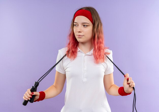 Junge sportliche Frau im Stirnband, die Springseil hält, das mit dem sicheren Lächeln auf Gesicht steht, das über lila Wand steht