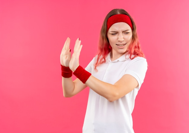 Junge sportliche Frau im Stirnband, die Verteidigungsgeste mit Hand mit angewidertem Ausdruck tut, der über rosa Wand steht