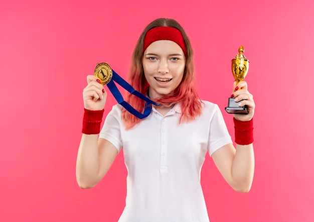 Junge sportliche Frau im Stirnband mit Goldmedaille um ihren Hals, die Trophäe mit Smilie auf Gesicht steht, das über rosa Wand steht