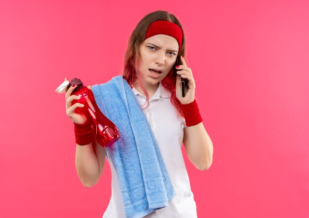 Junge sportliche Frau im Stirnband mit Handtuch auf Schulter, die Flasche Wasser hält, während am Telefon des Moniles spricht, das verwirrt und sehr besorgt steht, über rosa Wand steht