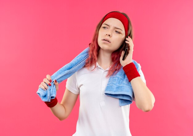 Junge sportliche Frau im Stirnband mit Handtuch auf Schulter, die mit ernstem Gesicht beiseite schaut, während auf Handy spricht, das über rosa Wand steht