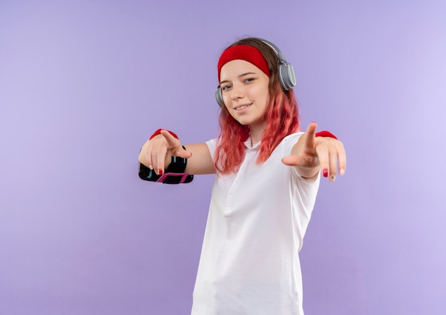 Junge sportliche Frau im Stirnband mit lächelnden Kopfhörern, die mit den Fingern zur Kamera zeigen, Training mit Smartphone-Armband, das über lila Wand steht