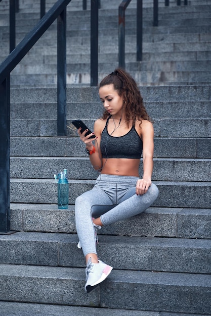 Junge sportliche frau in sportkleidung sitzt auf der treppe des städtischen stadions mit telefon in den händen und veröffentlicht ihre trainingsergebnisse Premium Fotos