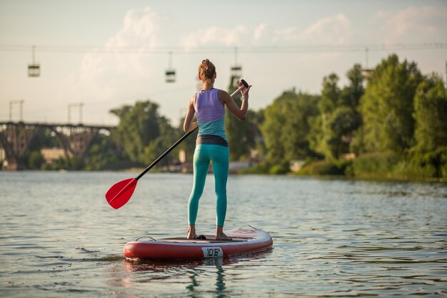 Junge sportliche Frau macht Fitness auf einem Brett mit einem Ruder auf einem See. Das Konzept eines gesunden Lebensstils. Sport. Yoga. Hobby