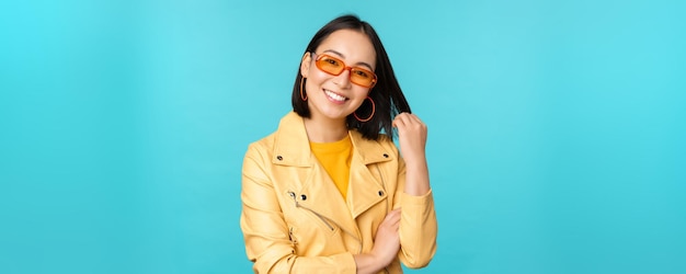 Junge stilvolle asiatische Frau mit Sonnenbrille lächelt, spielt mit ihrem Haarschnitt und sieht glücklich aus, posiert vor blauem Hintergrund