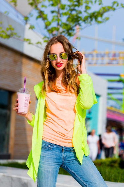 Junge stilvolle blonde Hipsterfrau in der kühlen Sonnenbrille mit Cocktail, der im Freien aufwirft