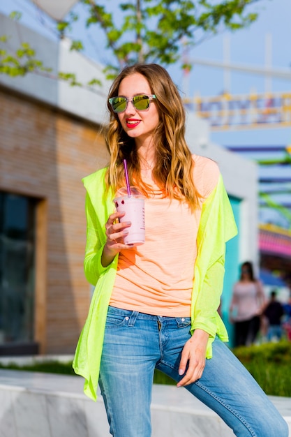 Junge stilvolle blonde Hipsterfrau in der kühlen Sonnenbrille mit Cocktail, der im Freien aufwirft