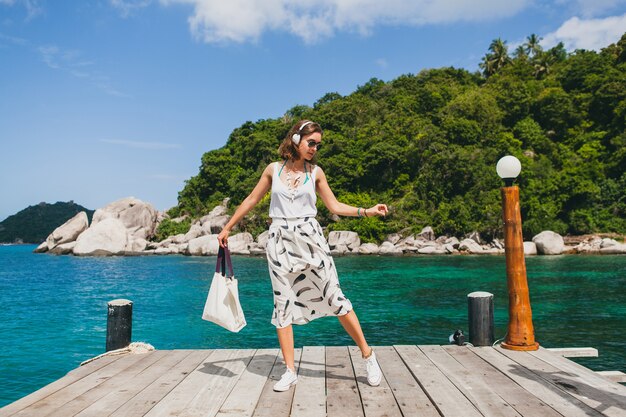 Junge stilvolle Frau, die auf Pier steht, zu Fuß geht, Musik auf Kopfhörern, Sommerkleidung, weißem Rock, Handtasche, azurblauem Wasser, Landschaftshintergrund, tropischer Lagune, Urlaub, Reisen in Asien hörend hört