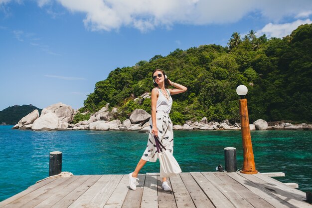 Junge stilvolle Frau, die auf Pier steht, zu Fuß geht, Musik auf Kopfhörern, Sommerkleidung, weißem Rock, Handtasche, azurblauem Wasser, Landschaftshintergrund, tropischer Lagune, Urlaub, Reisen in Asien hörend hört