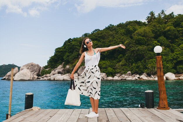 Junge stilvolle Frau, die auf Pier steht, zu Fuß geht, Musik auf Kopfhörern, Sommerkleidung, weißem Rock, Handtasche, azurblauem Wasser, Landschaftshintergrund, tropischer Lagune, Urlaub, Reisen in Asien hörend hört