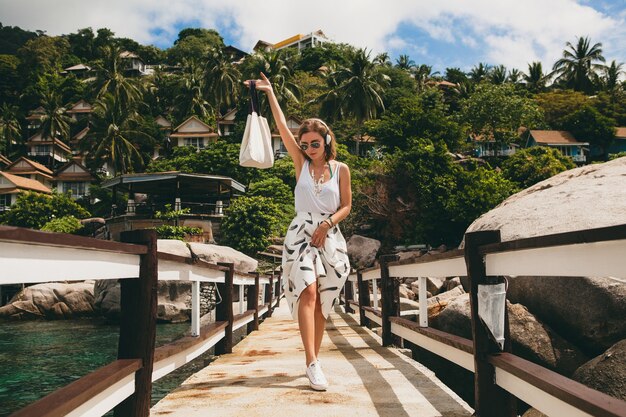 Junge stilvolle Frau, die auf Pier steht, zu Fuß geht, Musik auf Kopfhörern, Sommerkleidung, weißem Rock, Handtasche, azurblauem Wasser, Landschaftshintergrund, tropischer Lagune, Urlaub, Reisen in Asien hörend hört