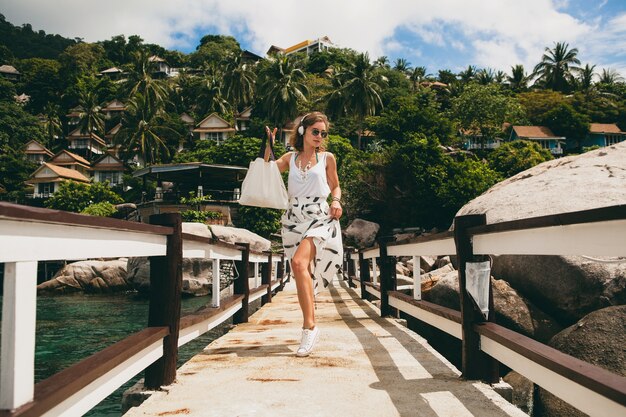 Junge stilvolle Frau, die auf Pier steht, zu Fuß geht, Musik auf Kopfhörern, Sommerkleidung, weißem Rock, Handtasche, azurblauem Wasser, Landschaftshintergrund, tropischer Lagune, Urlaub, Reisen in Asien hörend hört