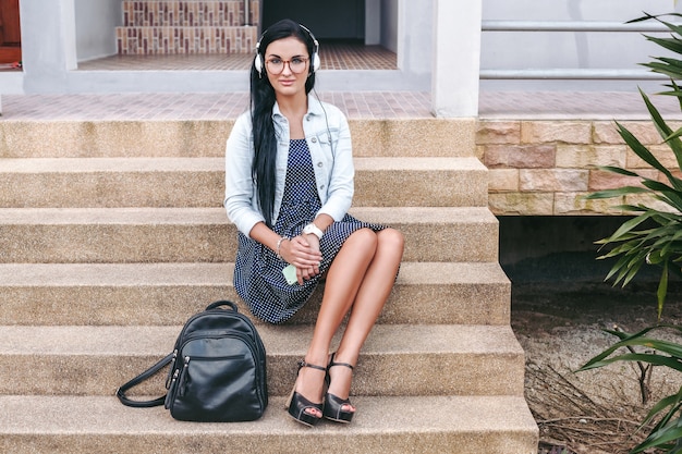 Junge stilvolle Frau, die auf Treppe mit Smartphone sitzt, Musik auf Kopfhörern, Rucksack hörend, lächelnd, glücklich, positive Stimmung, Sommerferien, Vintage-Denim-Stil