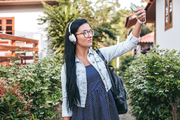Junge stilvolle Frau, die mit Smartphone geht, Musik auf Kopfhörern hörend, Foto, Vintage-Denim-Stil, Sommerferien machend