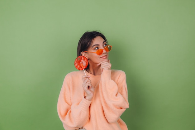 Junge stilvolle Frau im lässigen Pfirsichpullover und in den orangefarbenen Gläsern lokalisiert auf grüner Olivenwand mit nachdenklichem Blick des orangefarbenen Lutschers beiseite denkender Kopienraum