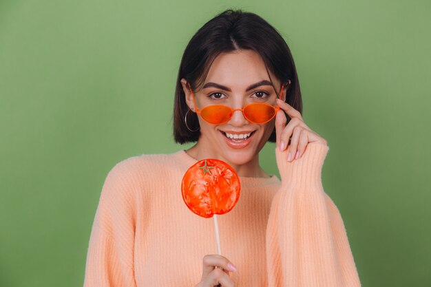 Junge stilvolle Frau im lässigen Pfirsichpullover und in den orangefarbenen Gläsern lokalisiert auf grüner Olivenwand mit positivem Lächelnkopienraum des orange Lutschers