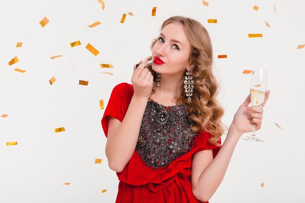 Junge stilvolle Frau im roten Abendkleid, das neues Jahr mit rotem Lippenstift feiert und Glas Champagner hält