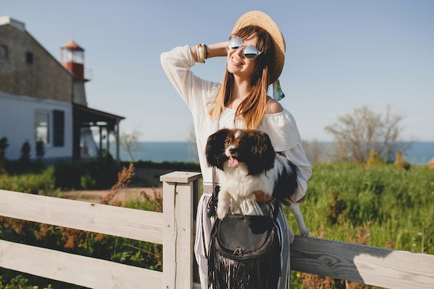 Junge stilvolle Frau in der Landschaft, einen Hund haltend, glückliche positive Stimmung, Sommer, Strohhut, böhmisches Stiloutfit, Sonnenbrille, lächelnd, glücklich, sonnig