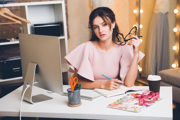 Junge stilvolle Frau in rosa Luxuskleid, die im Büro am Computer arbeitet