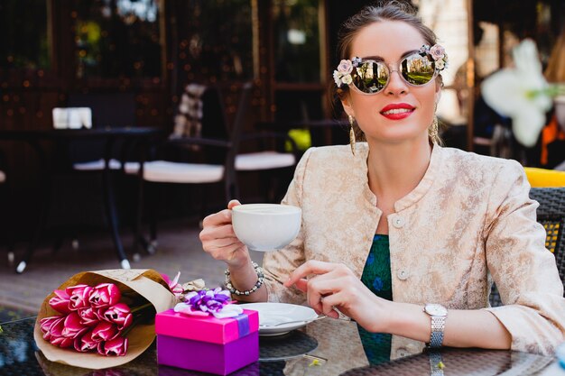 Junge stilvolle Frau, Mode-Sonnenbrille, die im Café sitzt und Tasse Cappuccino hält