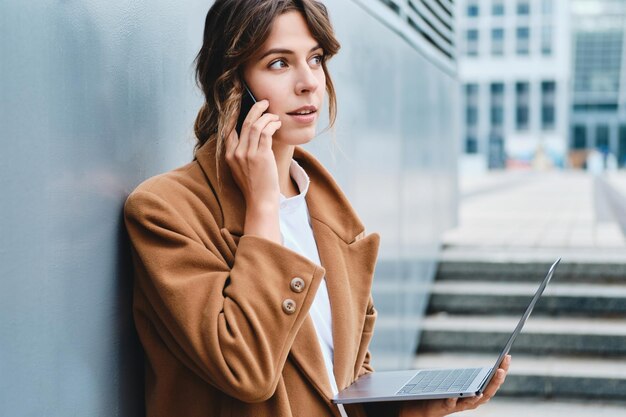 Junge, stilvolle Geschäftsfrau im Mantel, die nachdenklich am Handy spricht und auf der Straße der Stadt am Laptop arbeitet