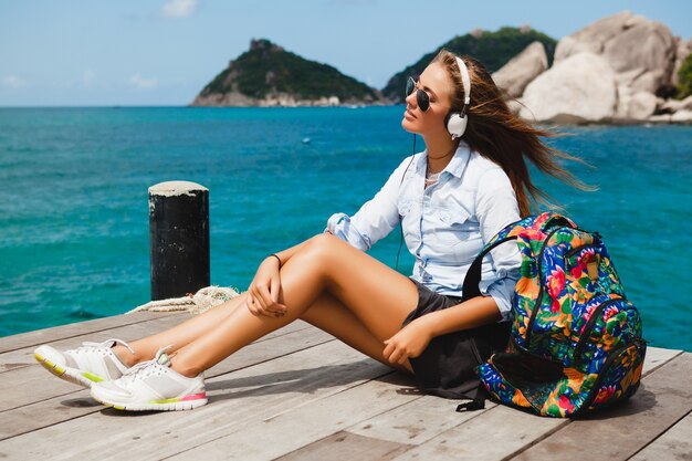 Junge stilvolle Hipster-Frau, die um die Welt reist, sitzt auf dem Pier, Fliegersonnenbrille, Kopfhörer, hört Musik, Urlaub, Rucksack, Jeanshemd, fröhliche, tropische Insellagune