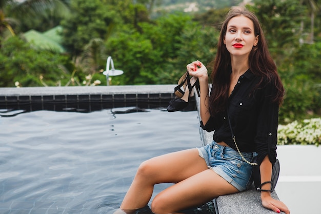 Kostenloses Foto junge stilvolle hipsterfrau, sitzend am pool, tropischer urlaub, sorglos, natürliche schönheit, schuhe in ihren händen haltend, rote lippen