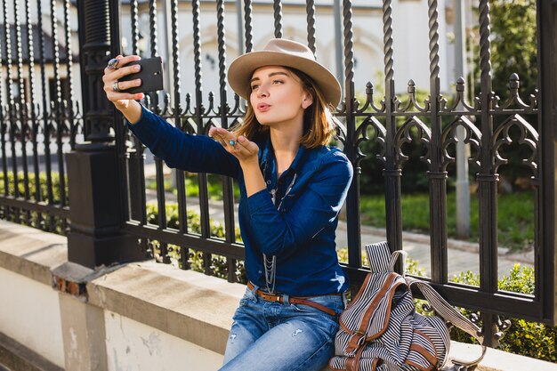 Junge stilvolle hübsche Frau, die einen Kuss durch ihr Telefon sendet, gekleidet in Jeanshemd und Jeans