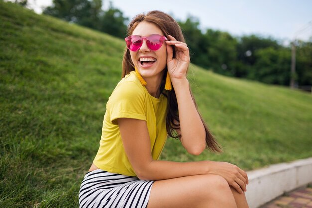 Junge stilvolle lachende Frau, die Spaß im Stadtpark hat, fröhliche Stimmung lächelt, gelbes Oberteil, gestreiften Minirock, rosa Sonnenbrille, Sommerart-Modetrend trägt