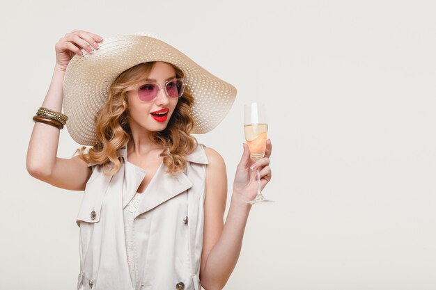Junge stilvolle lächelnde blonde Frau im großen Hut und in der Sonnenbrille, die Glas Champagner auf einer glücklichen Partei halten