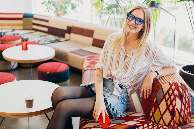 Junge stilvolle lächelnde Frau, die im Hipster-Café auf Sofa trägt Hemd und blaue Sonnenbrille sitzt