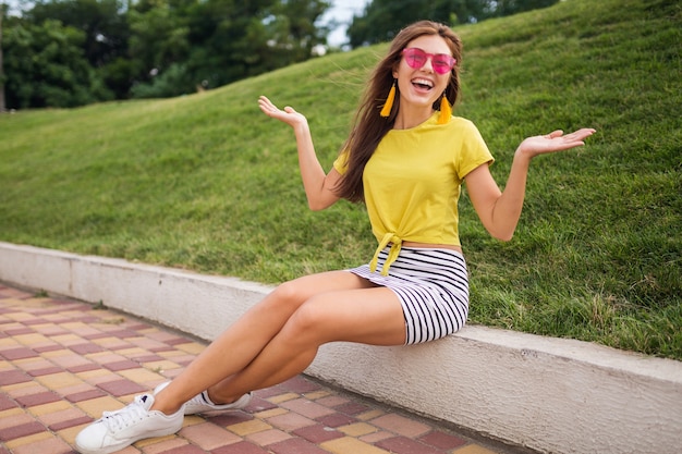 Junge stilvolle lächelnde Frau, die Spaß im Stadtpark hat, positiv, emotional, tragendes gelbes Oberteil, gestreiften Minirock, rosa Sonnenbrille, weiße Turnschuhe, Sommerart-Modetrend, glücklich, Händchen haltend