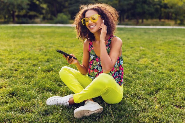 Junge stilvolle lächelnde schwarze Frau, die Smartphone verwendet, das Musik auf kabellosen Kopfhörern hört, die Spaß im Park haben, bunte Art der Sommermode, auf Gras sitzend, gelbe Sonnenbrille