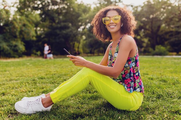 Junge stilvolle lächelnde schwarze Frau, die Smartphone verwendet, das Musik auf kabellosen Kopfhörern hört, die Spaß im Park haben, bunte Art der Sommermode, auf Gras sitzend, gelbe Sonnenbrille